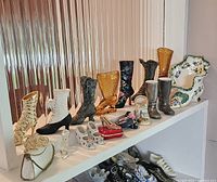 Shelf display of assorted miniature shoes and boots including a tall black boot, golden amber glass boot, white floral porcelain shoe, and more.