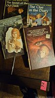 Four original Nancy Drew books laid out on a wooden surface showing covers including The Secret of the Old Clock, The Clue in the Diary, Mystery of the Tolling Bell, and Mystery of the Glowing Eye by Carolyn Keene.