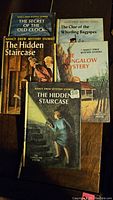 Five Nancy Drew books with visible covers on wooden surface, showing four titles and two editions of The Hidden Staircase.