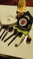 View of collection showing six pipes, one metal pipe with wooden ball handle, two ashtrays including Cardinal Golf Club logos, and ceramic animal figurine holding a pipe
