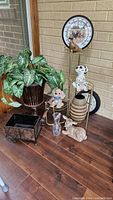 Photo of various decorative items arranged on a multi-tiered metal plant stand and on the floor, including ceramic figurines, a large clock, glass bottle, and a decorative metal container.