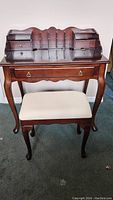 Front view of wooden secretary writing desk with multiple drawers and cream upholstered stool underneath