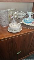 Photo showing clear etched cut-glass vase, footed glass bowls with floral designs, blue and white colored glass bowl with handle, and floral patterned china teacup with saucer and spoon on wooden cabinet.