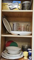 Shelves containing a metal container, sharpening stone, clear glass measuring cups, various ceramic plates, bowls, and egg cups.