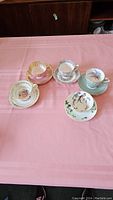 Five vintage teacups and matching saucers arranged on pink tablecloth, showing variety of patterns and designs