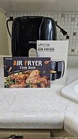 Black VPCOK Air Fryer with manual and cookbook displayed on kitchen counter, showing front view and brand name.
