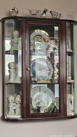 Wall mounted curio cabinet with dark wood frame, curved glass front and sides, mirrored back, containing decorative plates and figurines.