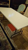 White folding utility table with black metal folding legs, approx 6 feet long, shown indoors near furniture.