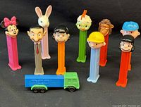 Eight PEZ candy dispensers standing upright with colorful character tops and one PEZ toy truck in front.