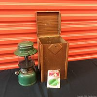 Green Coleman lantern with glass globe, Coleman label, and metal frame, next to a tall wooden box with diamond-shaped wooden decoration on the front, and a sealed packet containing an extra mantle placed in front.