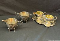 Photo showing two silver plated cream and sugar bowl sets, one set includes a small ornate tray. Each piece has decorative handles and engraved detailing.