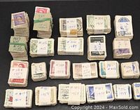 Photo showing 25 bundles of mostly used postage stamps wrapped in paper strips. Each paper strip hand-labeled with catalogue numbers and years ranging from 1950 to 1964. Stamps mainly Canadian.