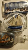 Photos showing three different ships in glass bottles. Top bottle has a white ocean liner ship, middle bottle a two-masted schooner, lower bottle a sailing ship with brown hull.