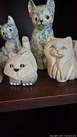 Five cat figurines arranged on a wooden shelf, showing two carved stone cats and two decoupage cats in front, plus a textured cat figurine.