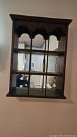 Full view of the dark wood curio cabinet mounted on a wall showing the mirrored back and three shelves with vertical decorative dividers.