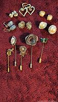 Photo shows all six pairs of earrings and five stick pins arranged on a fabric background. Earrings include designs with clear stones, open hearts, grey stones, and pearls. Stick pins include diverse decorative tops such as a rose, butterfly, and floral motifs.