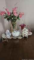 Glass vase with pink flowers set in a gold flowing brand and leaf designed stand. Several assorted china collectibles next to the vase including floral covered dish, trinket box, and small china figurines. Also visible is a round gold frame with mirror.