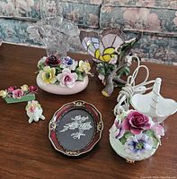 Photo showing multiple china flower arrangements, a stained glass butterfly light, a framed floral embroidery, and various ceramic flower decorations on wooden table.