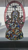 Front view of oval stained glass peacock window hanging suspended by chain, showing peacock motif and rose floral border