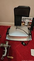 Photo showing the Heritage Rock Plus reversible grill and griddle on the floor, with electric warming pan on top and the manual metal meat grinder beside it. A wood cutting board and the Heritage Rock Plus instruction booklet are also visible.