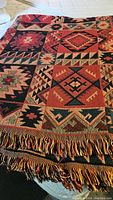 Full view of colorful woven blanket showing large geometric and tribal patterns and fringed edge in multiple earth tone colors.