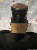 Photo showing the coffee press resting on its packaging box. The coffee press has a black finish with a wood-tone top, depicted from a top angle.