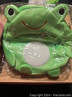 Front view of new small green frog plush backpack in plastic packaging, showing frog face with eyes and smile and white belly patch.