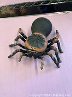 Large rubber spider figurine placed on a pink surface with black backing, showing detailed legs and textured body.