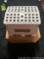 White folding step stool fully opened on a box, showing the top with holes and a grid pattern.