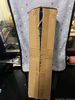 Closed cardboard box containing a small artificial Christmas tree, showing the box from the front with some damage and tape remnants.