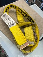 Yellowfeet branded yellow truck strap with black 'U.S. CA' lettering, metal ratchet, and hook hardware laid on beige surface.