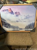 Photo of a framed wall painting showing a mountain landscape covered in snow and clouds, with pine trees and a lake below.