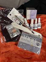 Wellness and beauty product assortment laid on red fabric: nail care polishes and gel system, lip repair balm, eye shadow containers, makeup brush in packaging, dark fabric item (ice pack cover), boxed skin care product