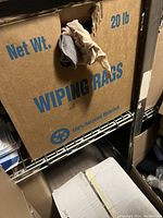 Photo showing a brown cardboard box labeled 'Wiping Rags Net Wt. 20 lb' with some cleaning rags protruding from a hole in the box. The box sits on a metal shelf among other boxes and items.
