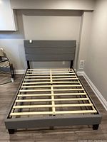 Full frontal view of gray upholstered bed frame with horizontal stitched padded headboard and wooden slat base, black legs.