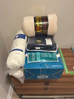 Photo showing Matfix Home fleece blanket, Ikea mattress pad, blue dark bedding item with Glow lotion package, and boxed comforter set stacked on wooden dresser.
