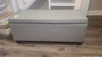 Front view of grey fabric storage bench with black block feet on wooden floor.