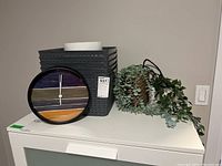 Photo showing a 10-inch battery-operated wall clock with a black frame, abstract multicolor face, two faux plants (one hanging in wicker pot and one in stone pot), three gray plastic storage baskets stacked, and a white waste bin.