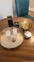 Photo showing a gold-tone metal decorative bowl, two black and gold rectangular storage tins, two clear frosted glass tealight holders, a large round hammered metal tray, and a small glass hourglass timer on a wood table.