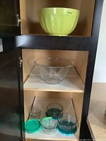 Top shelf of cabinet with light green ceramic mixing bowl, below a clear glass bowl, and bottom shelf with multiple glass storage bowls with teal lids and one small glass bowl without lid.