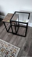 Two nested end tables, one with wood top and one with glass top, both on steel frames.
