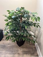 Front view of artificial plant with green leaves in dark brown wicker planter on wood floor against cream walls.