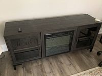 Full front view of the electric fireplace cabinet showing dark composite wood construction, two glass door compartments, and central fireplace unit. Remote control placed on top of cabinet.