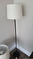 Full view of the black metal floor lamp standing in a corner, showing the white cylindrical lampshade and base.