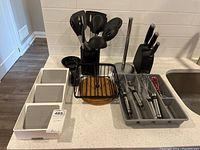 Photo showing assortment of kitchen utensils in black utensil holder, Kuradori knife set with black block, white multi-compartment serviette holder, black wire basket and kitchen tools in grey tray on kitchen counter.