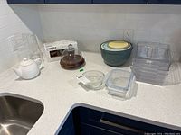 Overview of all kitchenware items on white countertop including teapot, glass pitcher, butter dish, nesting bowls, plastic and glass storage containers, and boxed Ikea canister set.