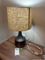 Photo showing lamp front with beige fabric shade and matte black ceramic base with metallic accent, power cord visible.