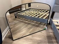 Front view of wall mirror showing the slight arch and black metal frame, leaning against a bed frame.