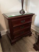 Front and side view of the wooden nightstand showing three drawers with metal pulls and removable glass top with lamp on it.