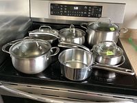 Photo of 6 stainless steel Lagostina pots and pans with 5 glass lids on stove, showing entire set and extra T-fal pan.